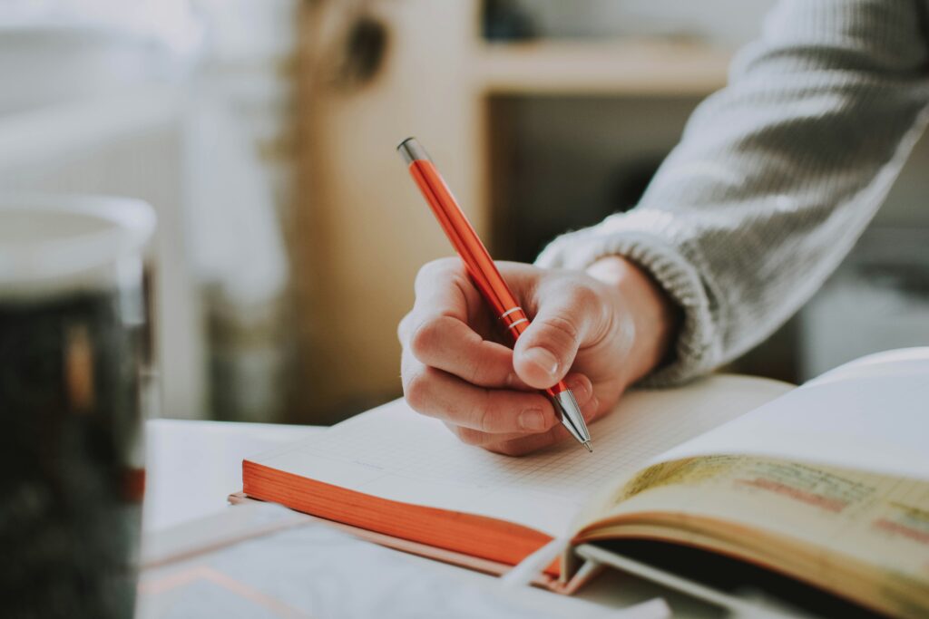 A close-up of a hand holding a red pen while writing in a notebook, with a warm and soft background setting.
